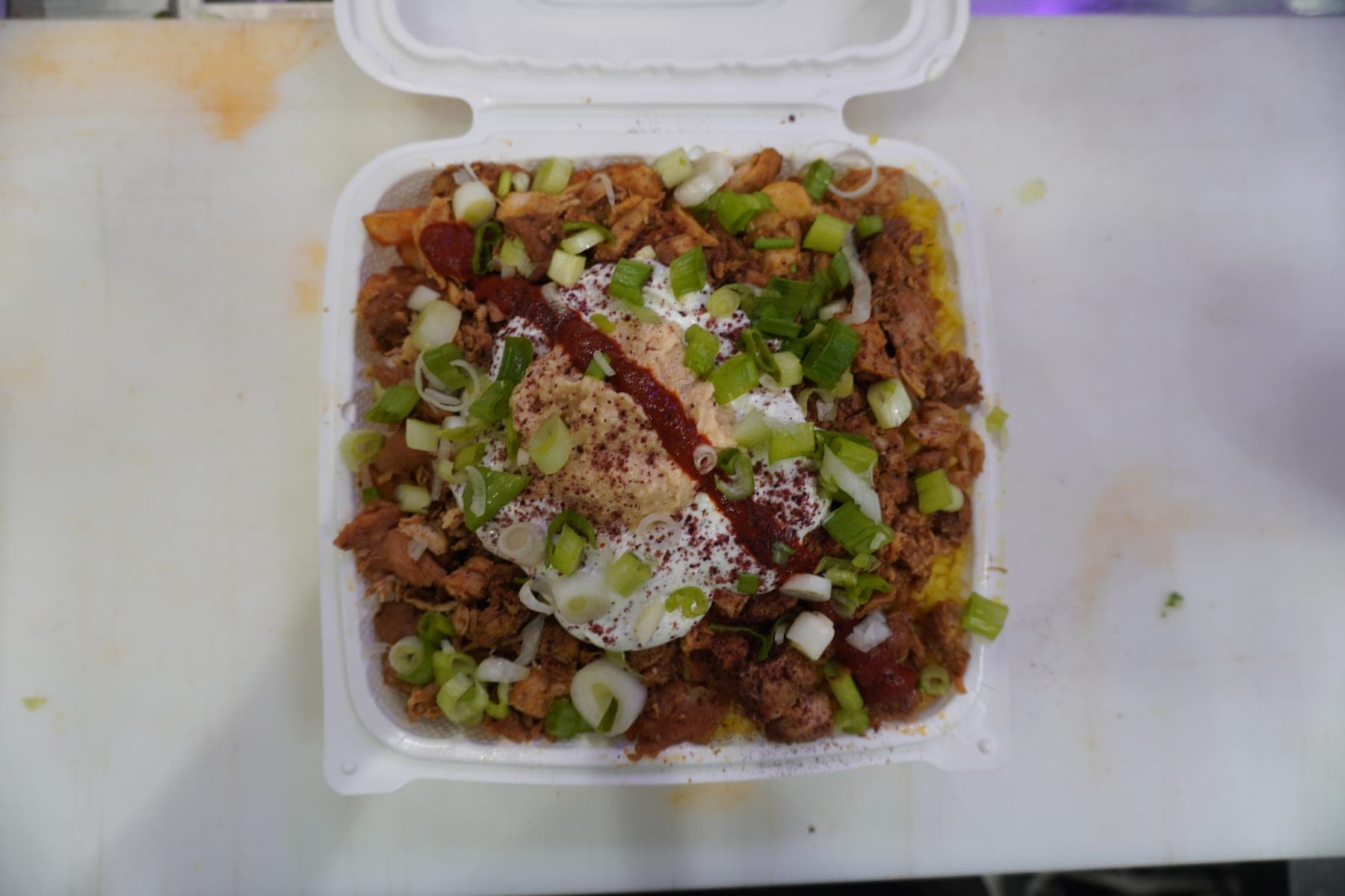 A chicken bowl in the box, with hummus, hot sauce, sumac and green onion over the top